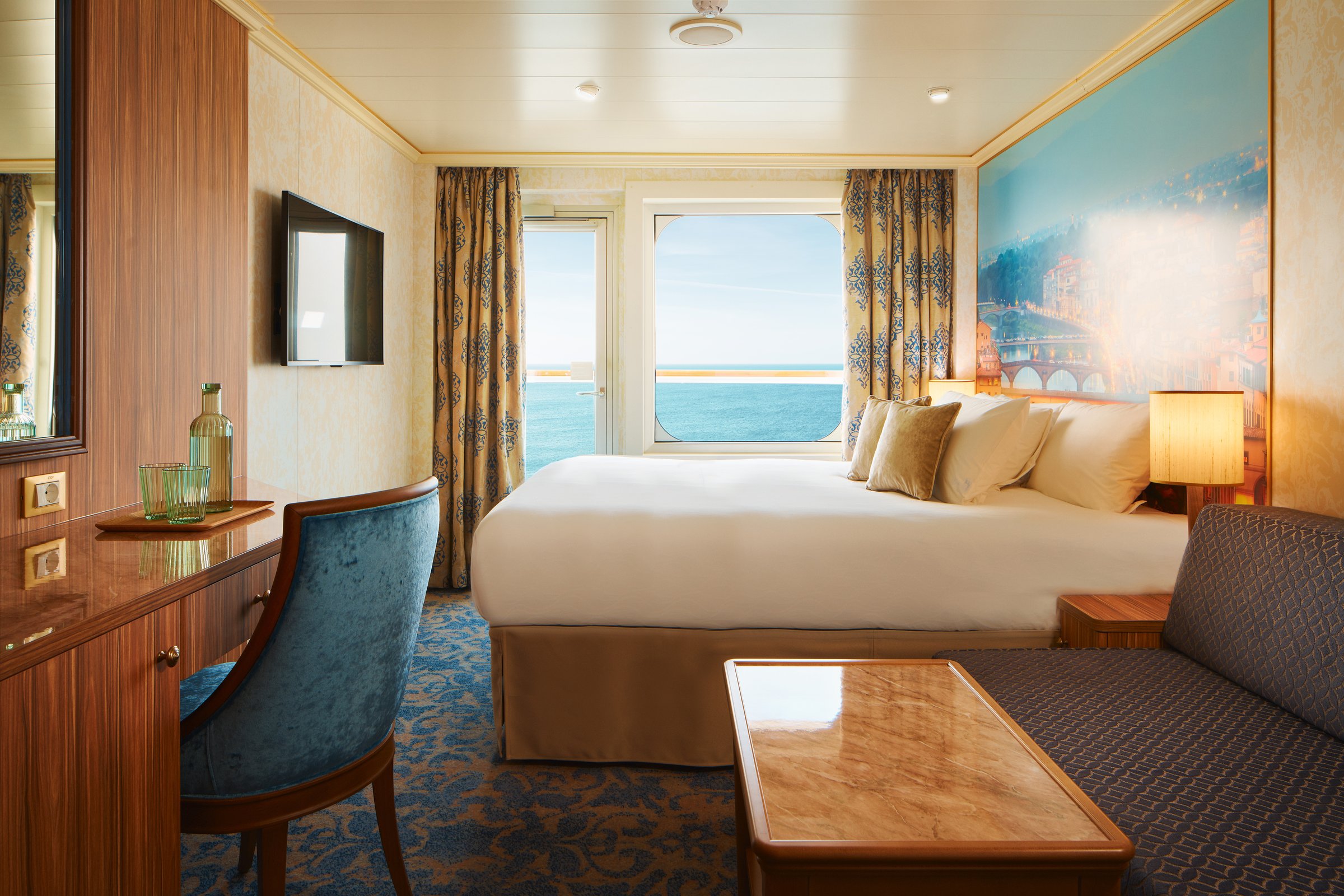 Carnival Firenze balcony stateroom with a large bed, sofa and a blue Italian-scene mural under daylight from the balcony door