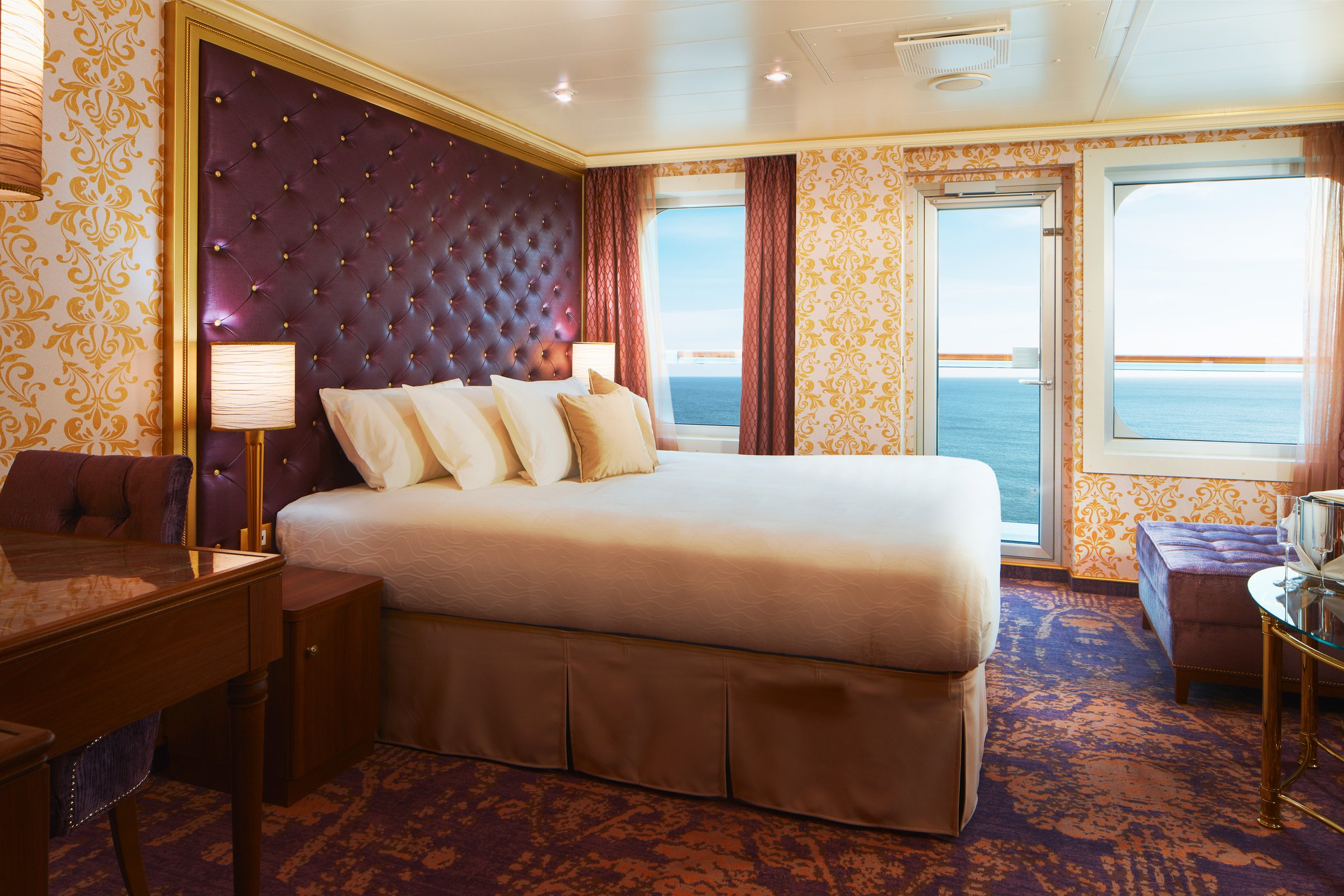 Carnival Firenze suite with a tufted purple headboard, gold damask wallpaper and balcony doors to the sea