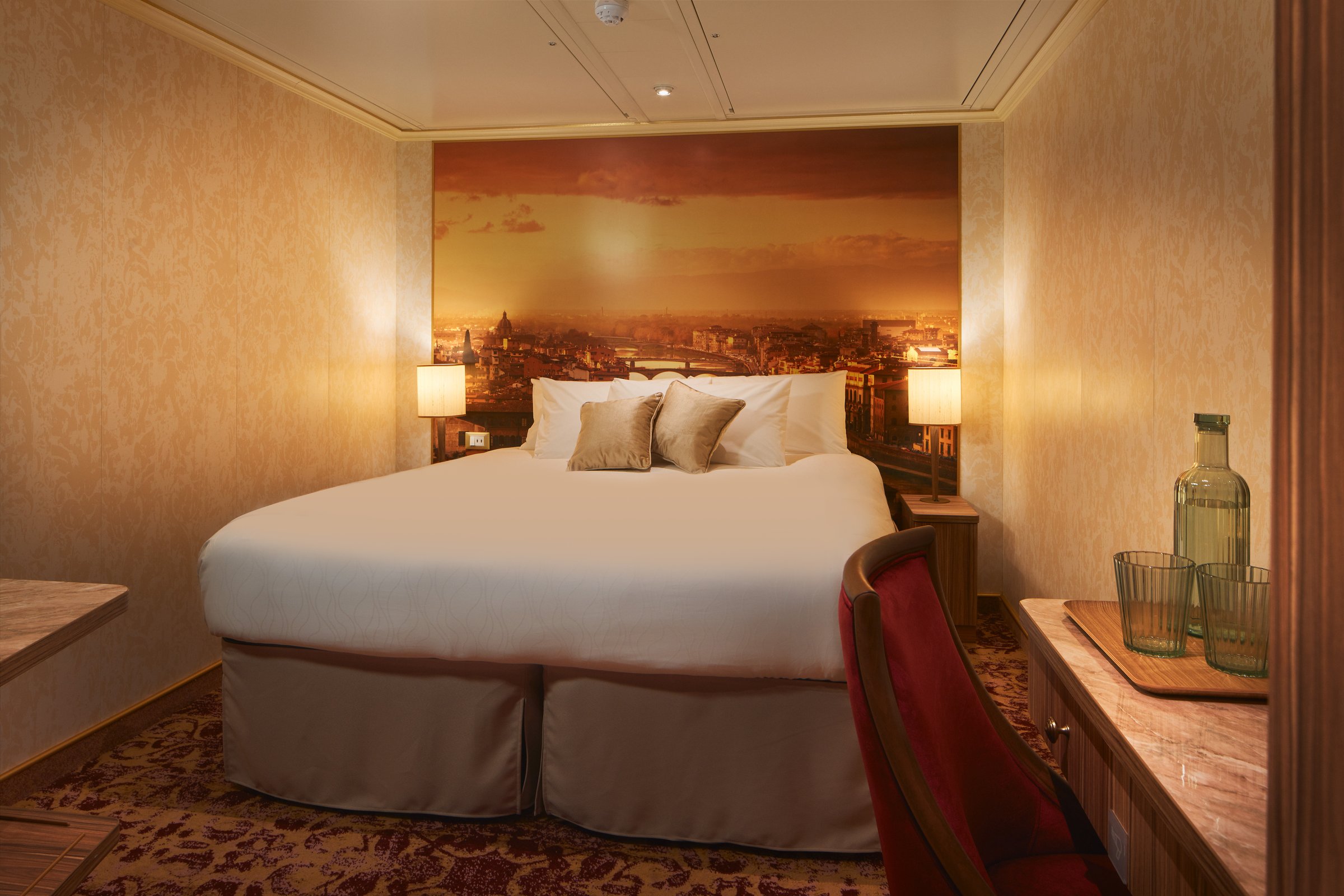 Carnival Firenze interior stateroom with a double bed, warm lamps and a sunset city mural
