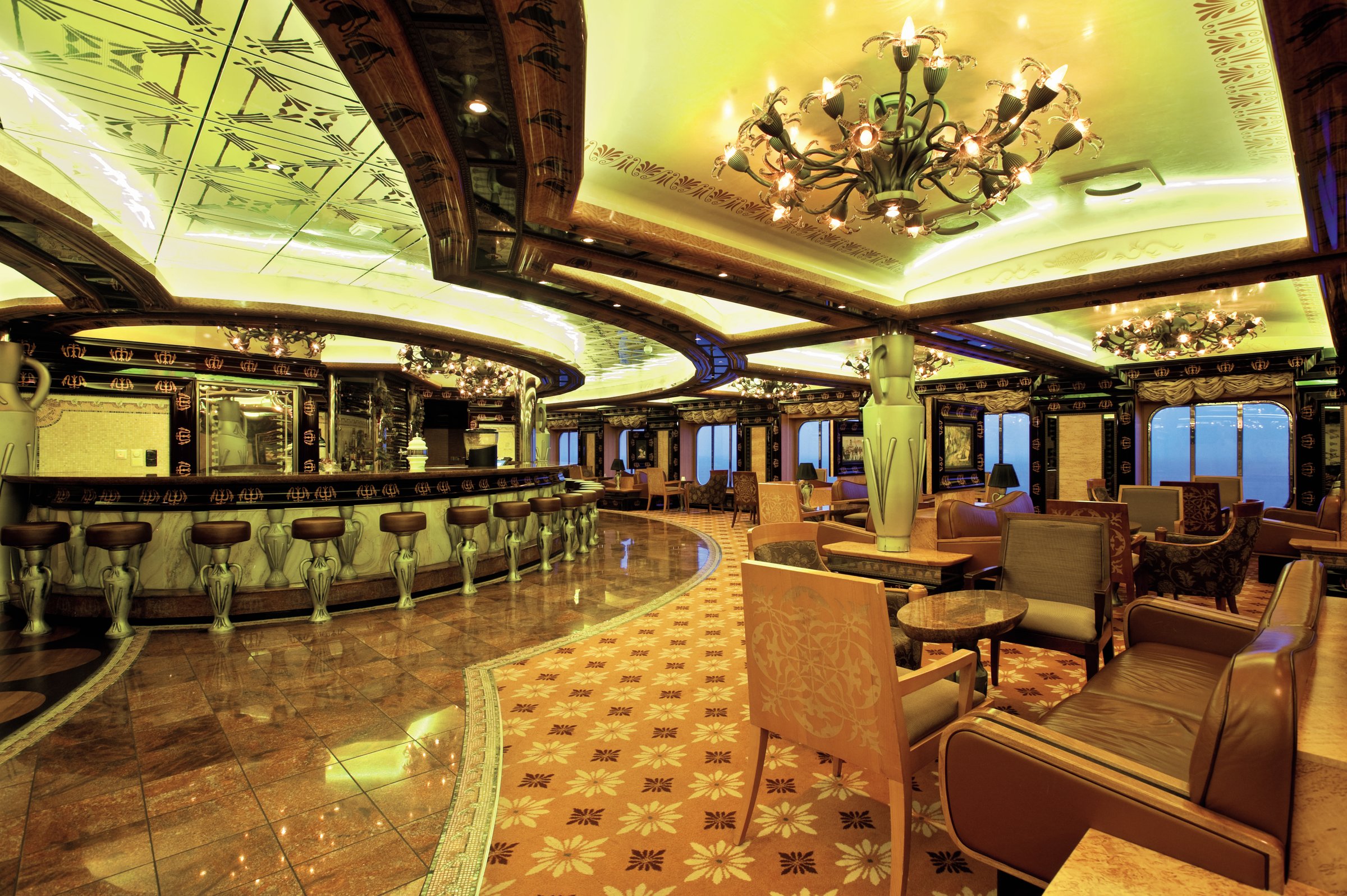 Carnival Legend Atlantis lounge with a long bar, upholstered armchairs, a chandelier and a patterned floor