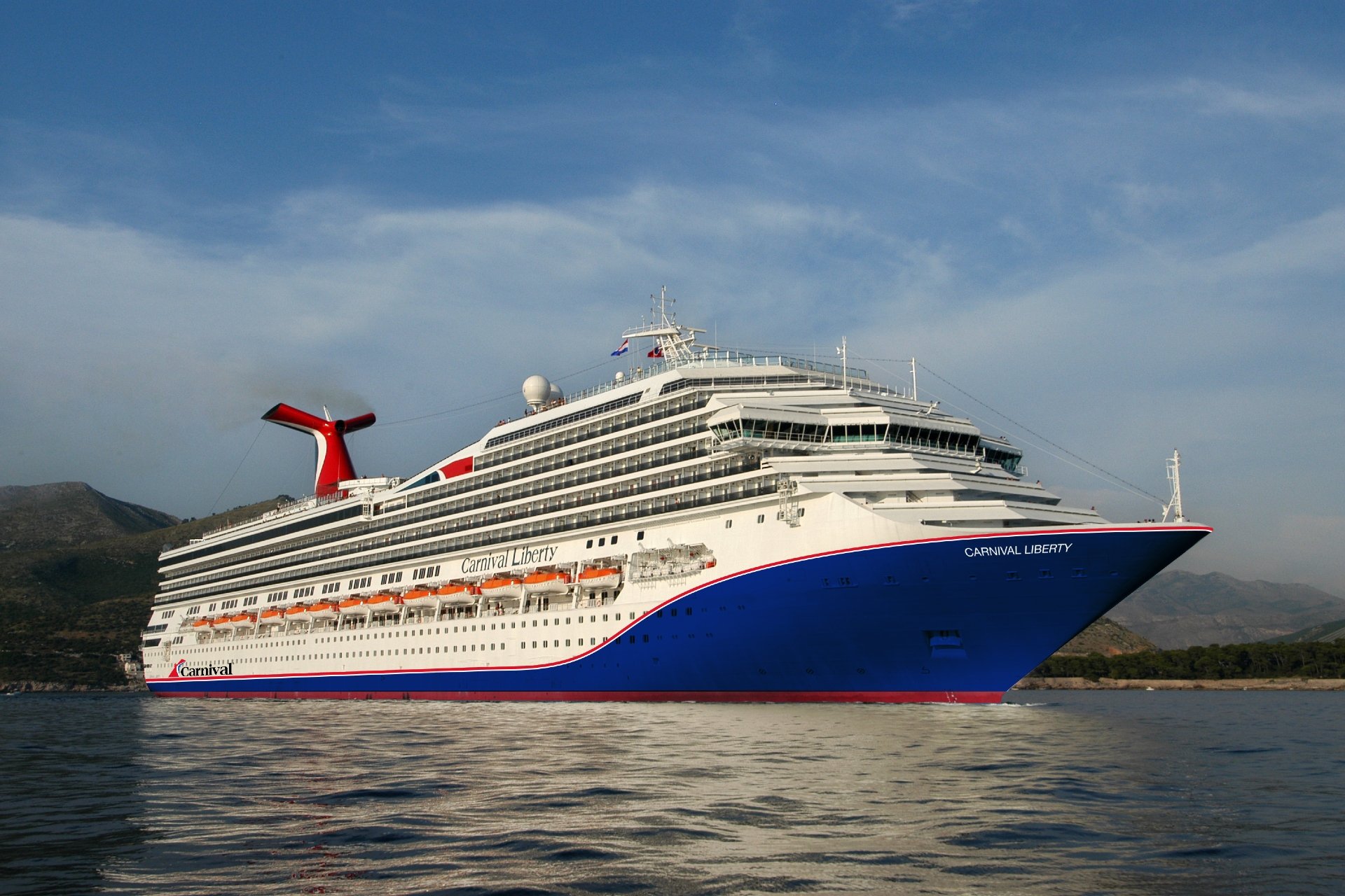Carnival Liberty at sea, bow three-quarter view with blue hull and red funnel against coastal hills