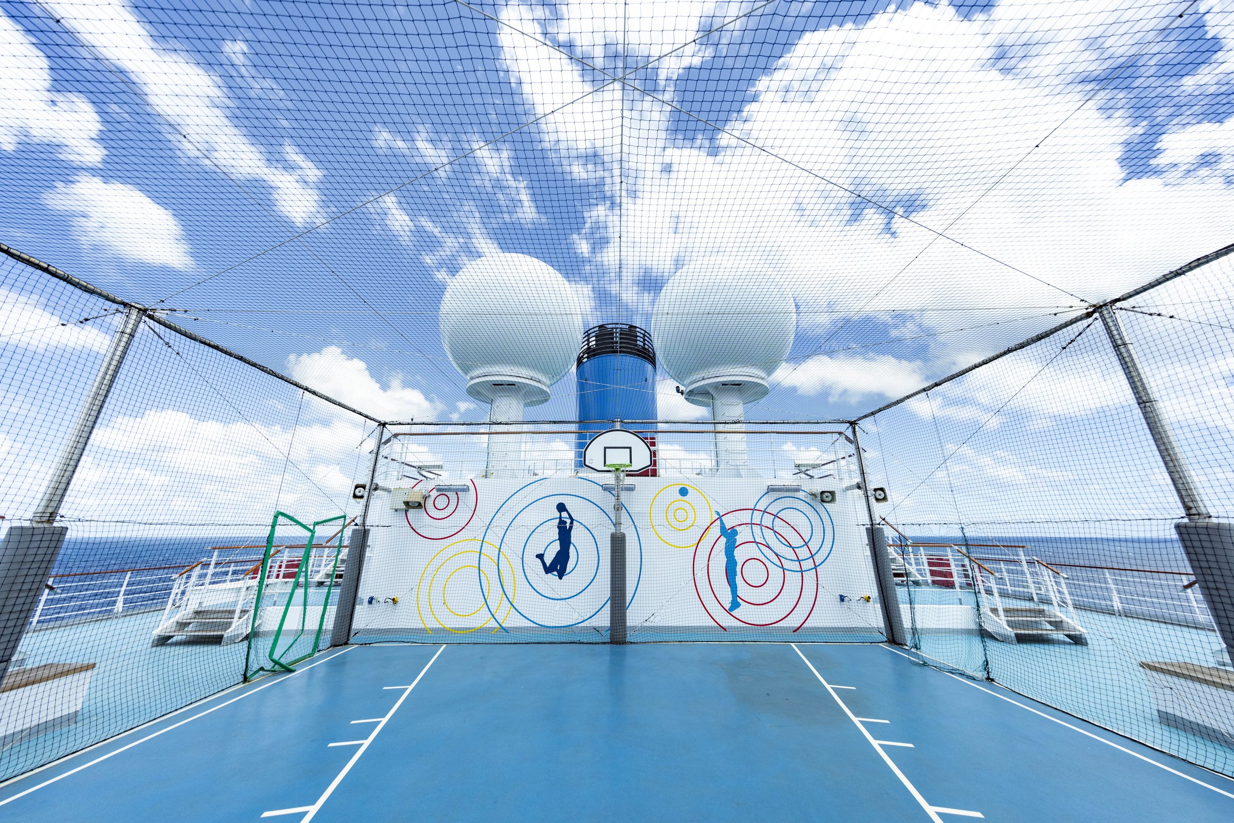 Carnival Luminosa sports court with a blue floor, basketball hoop and netting under a cloudy sky