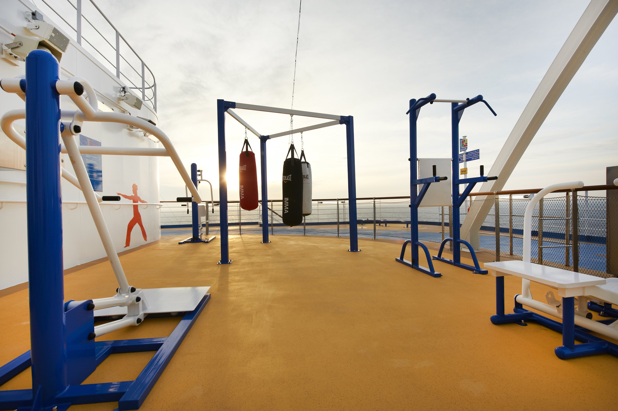 Carnival Magic sports deck with boxing bags, blue fitness stations and an orange rubberised floor in warm low sun