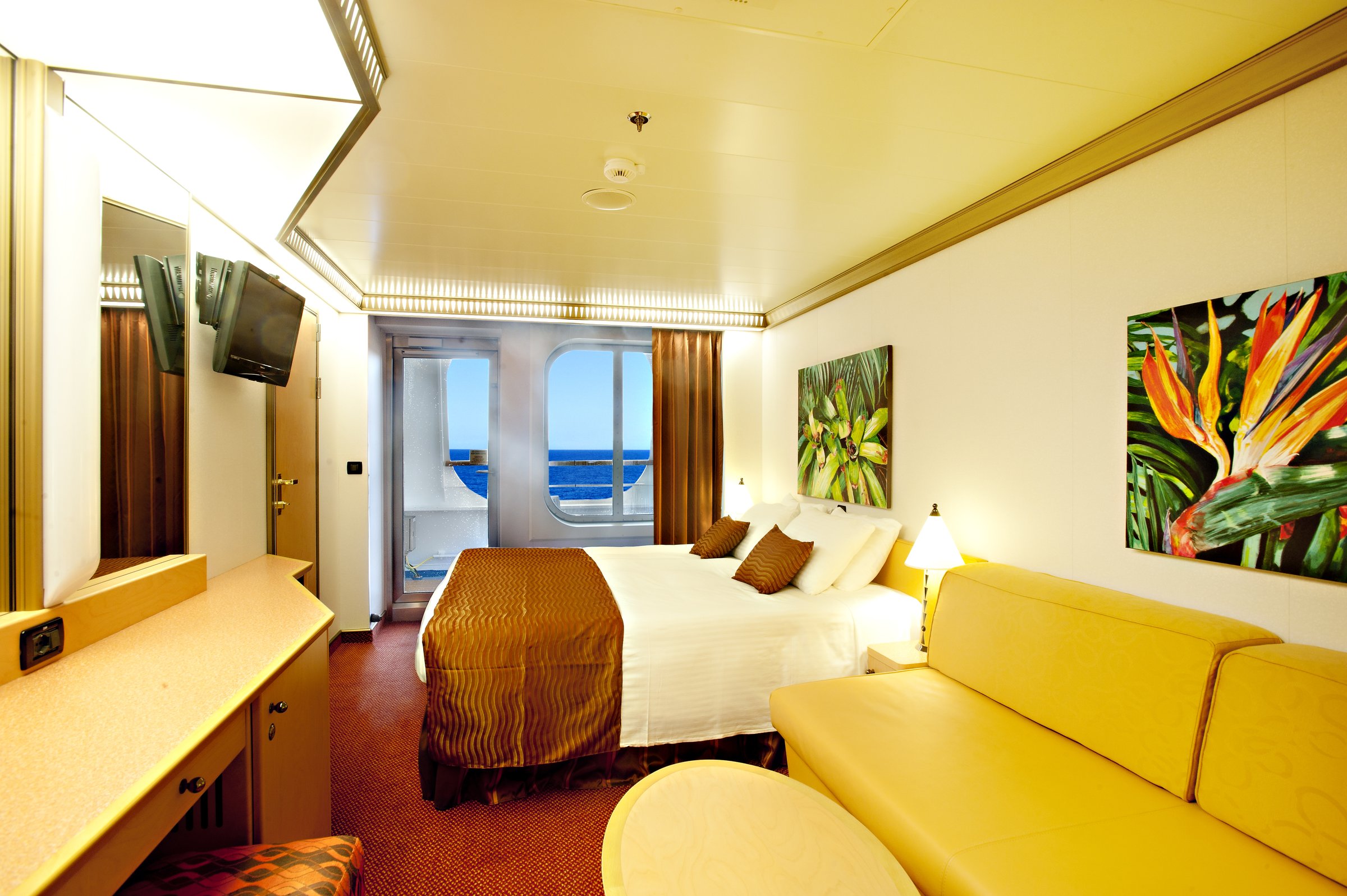 Carnival Magic balcony stateroom with a king bed, sofa, tropical artwork and a sea view through the balcony door