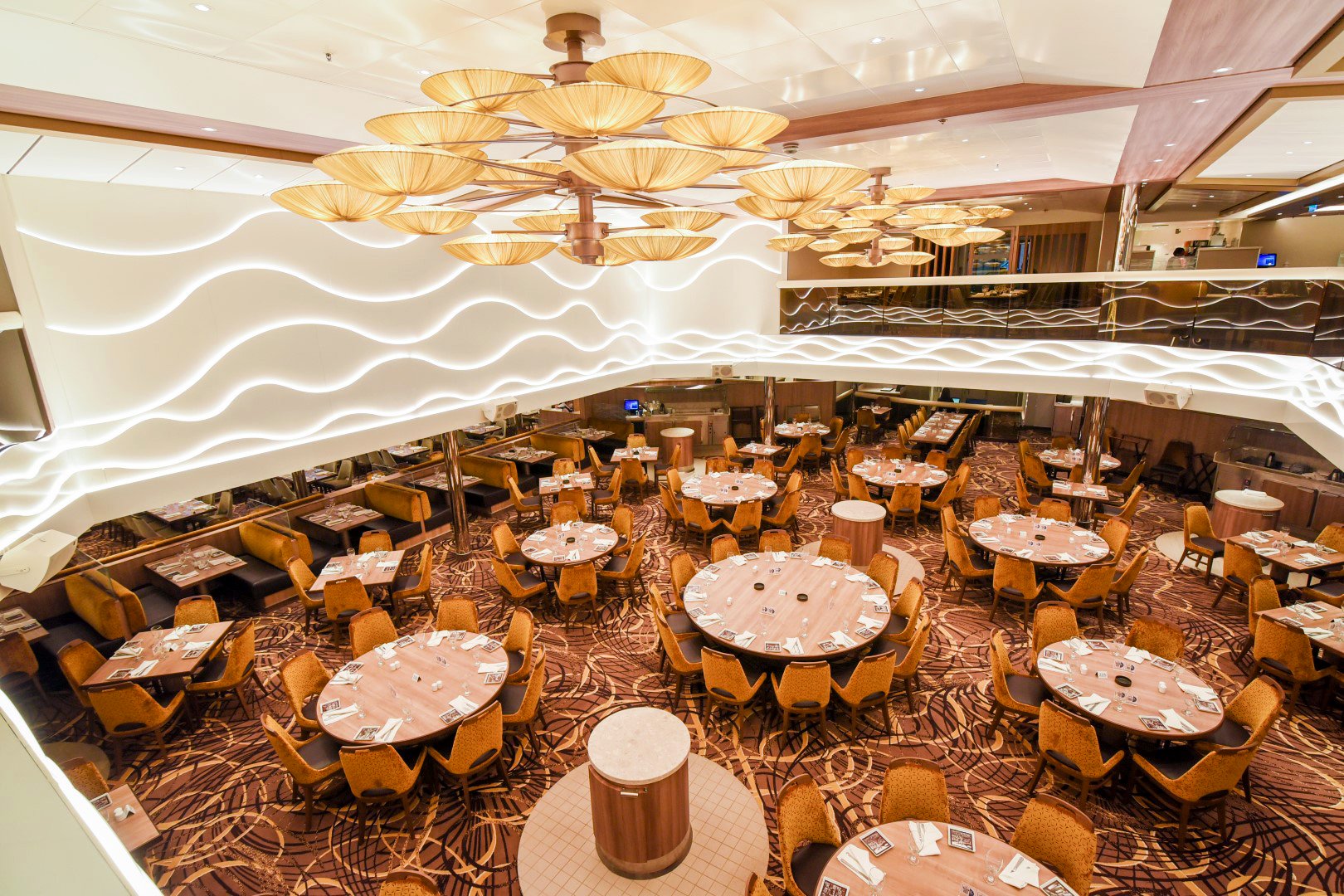 Carnival Radiance main dining room with round tables, gold chairs, wave-patterned walls and petal chandeliers