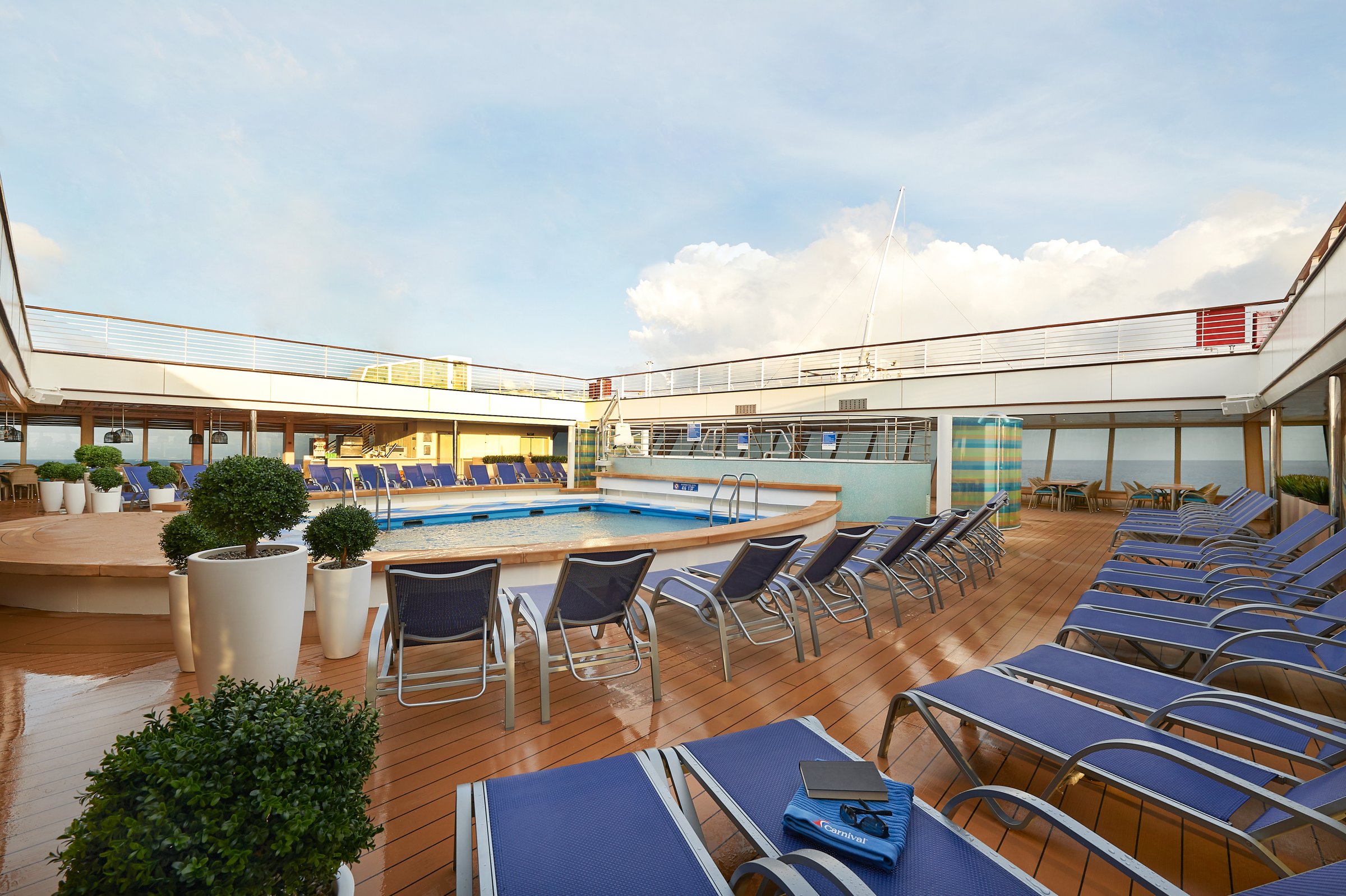 Carnival Sunrise aft pool with blue loungers, topiary planters and a bright cloudy sky