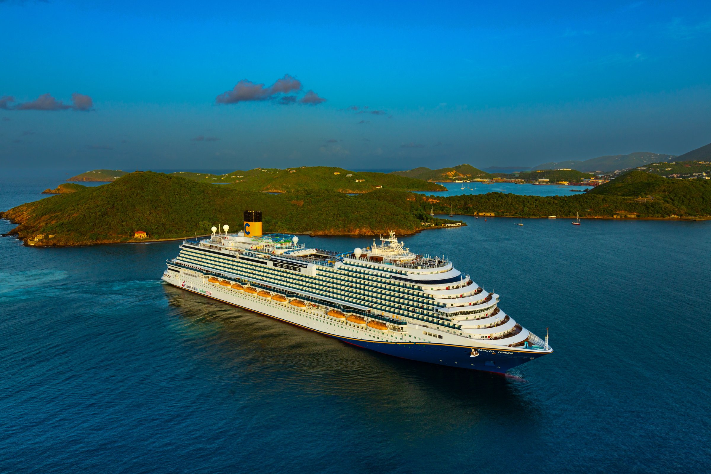 Aerial of Carnival Venezia cruising past green islands in a deep-blue bay on a sunny day