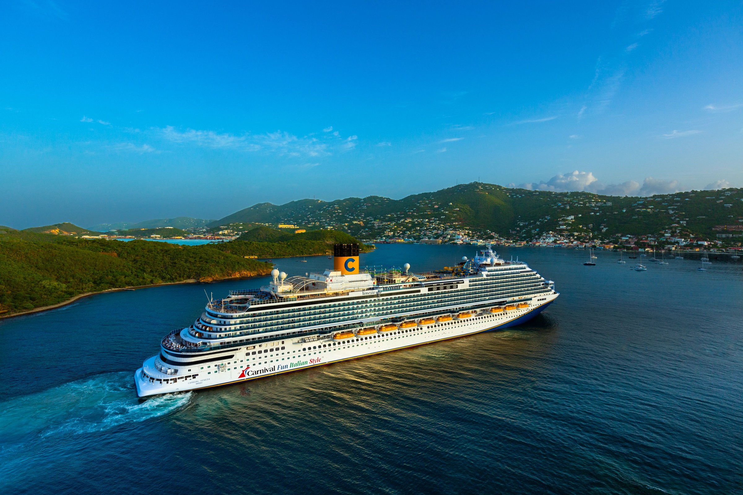 Aerial of Carnival Venezia sailing into a green island harbor under a clear blue sky