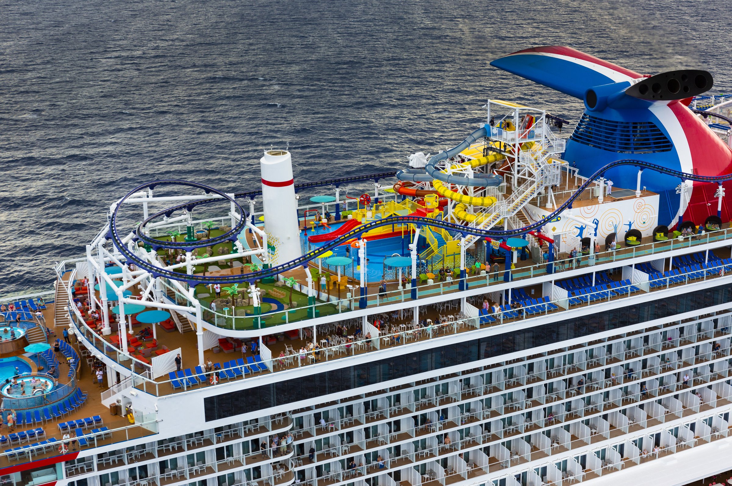 Aerial of the Mardi Gras top decks with the funnel, BOLT coaster and WaterWorks above a blue sea
