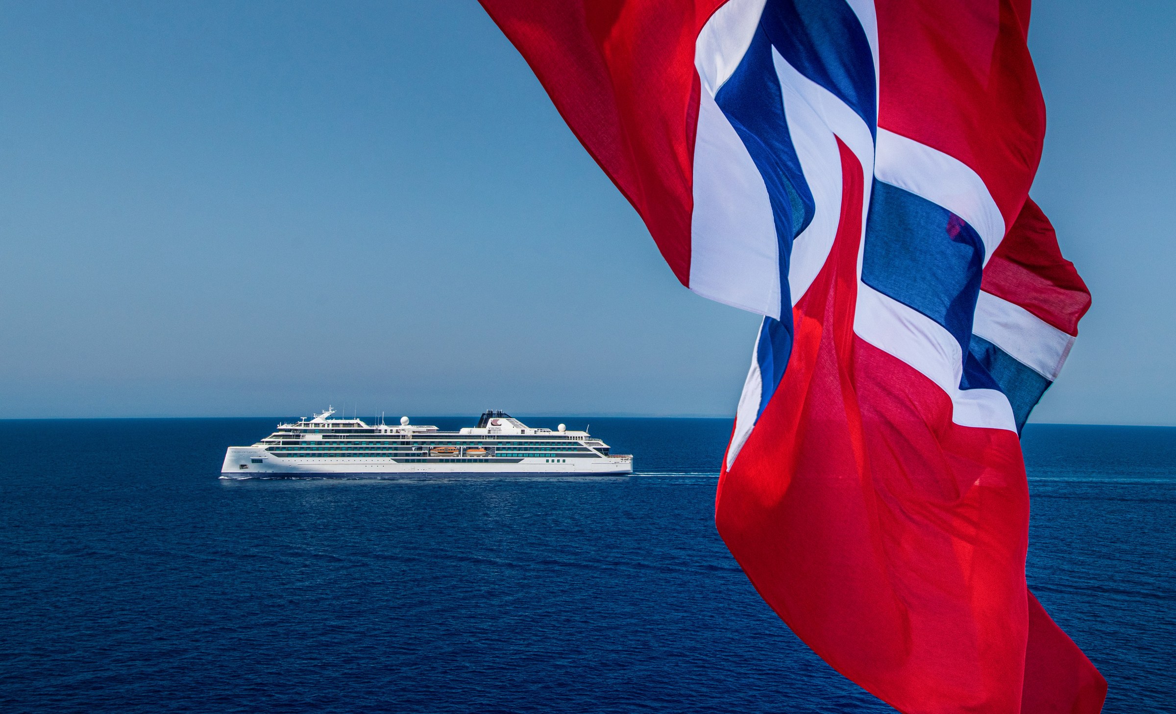 Viking Octantis sailing on a bright blue sea beyond a Norwegian flag in the foreground