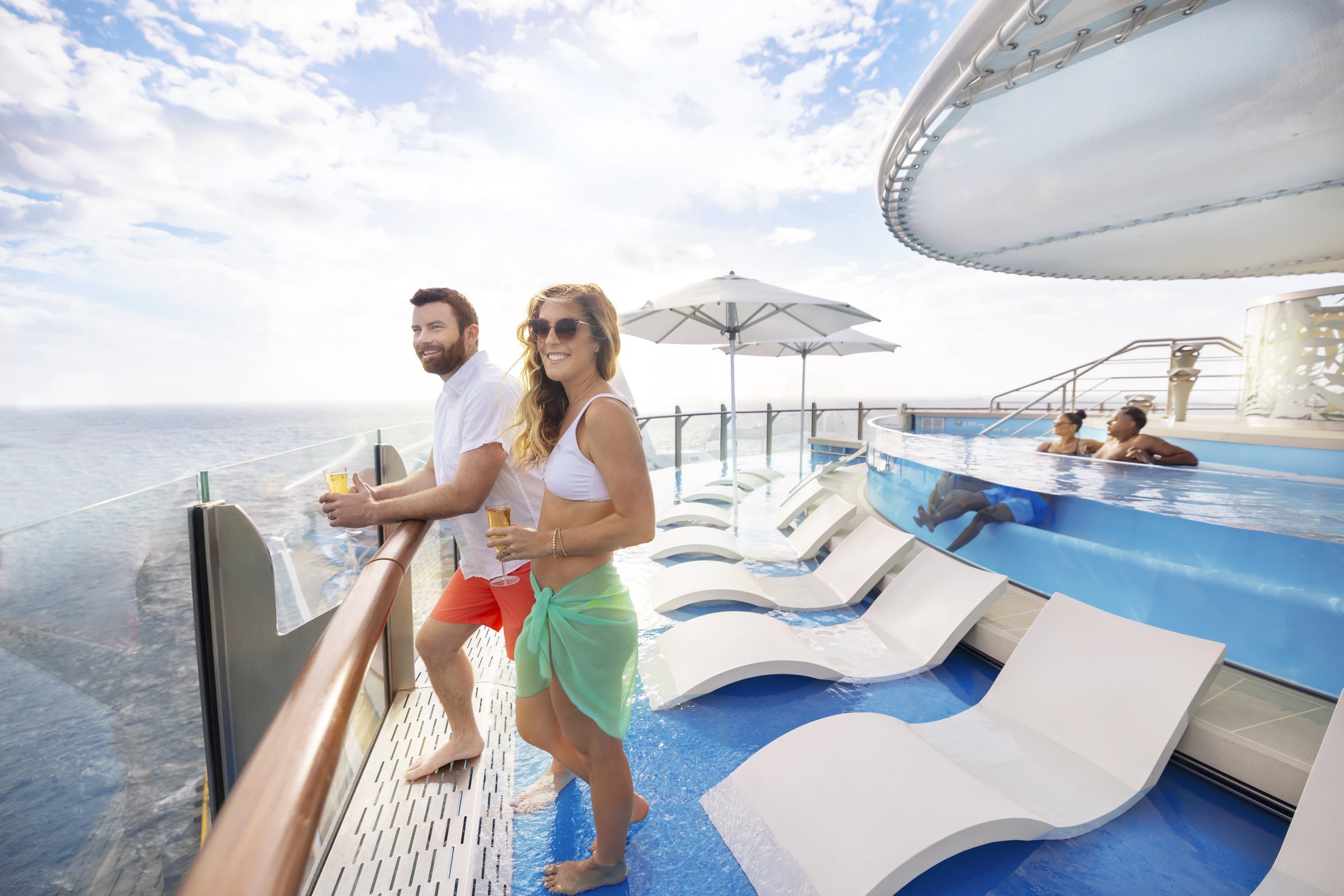 Couple with drinks at the rail beside the Suite Sun Deck pool aboard Wonder of the Seas, sunny sea view